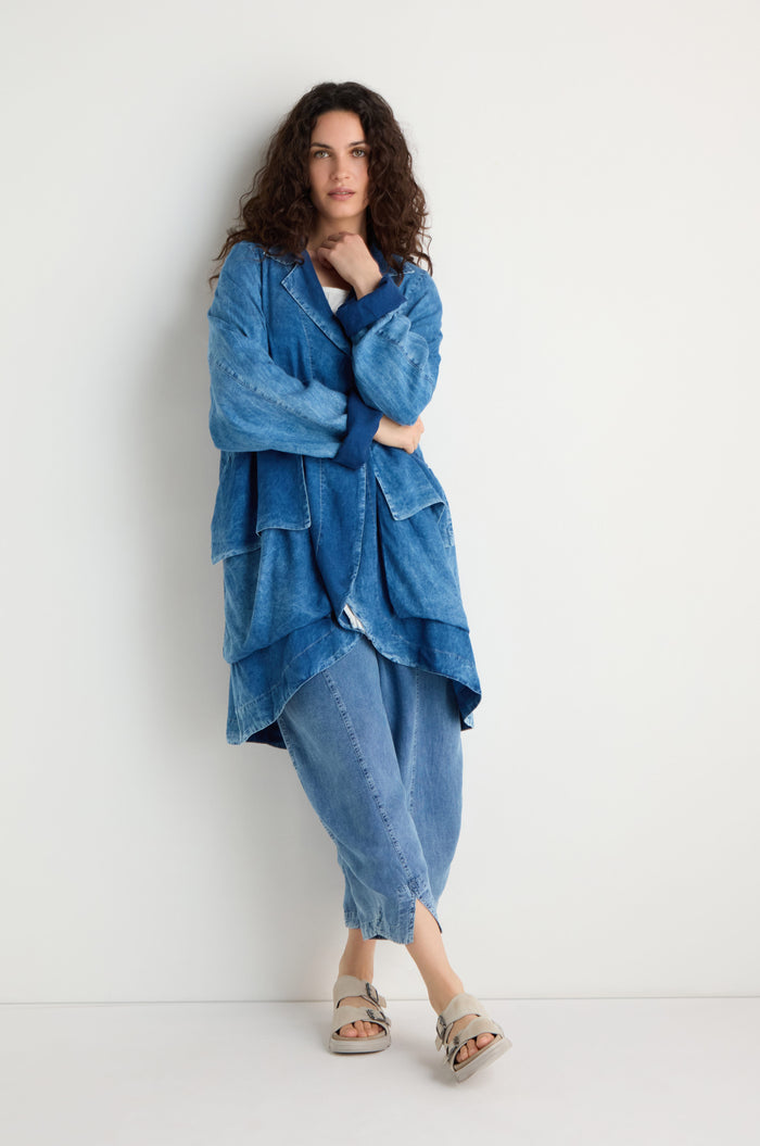 A person with long curly hair wears a Lyocell Utility Jacket in blue with matching pants and beige sandals, standing against a plain white wall.