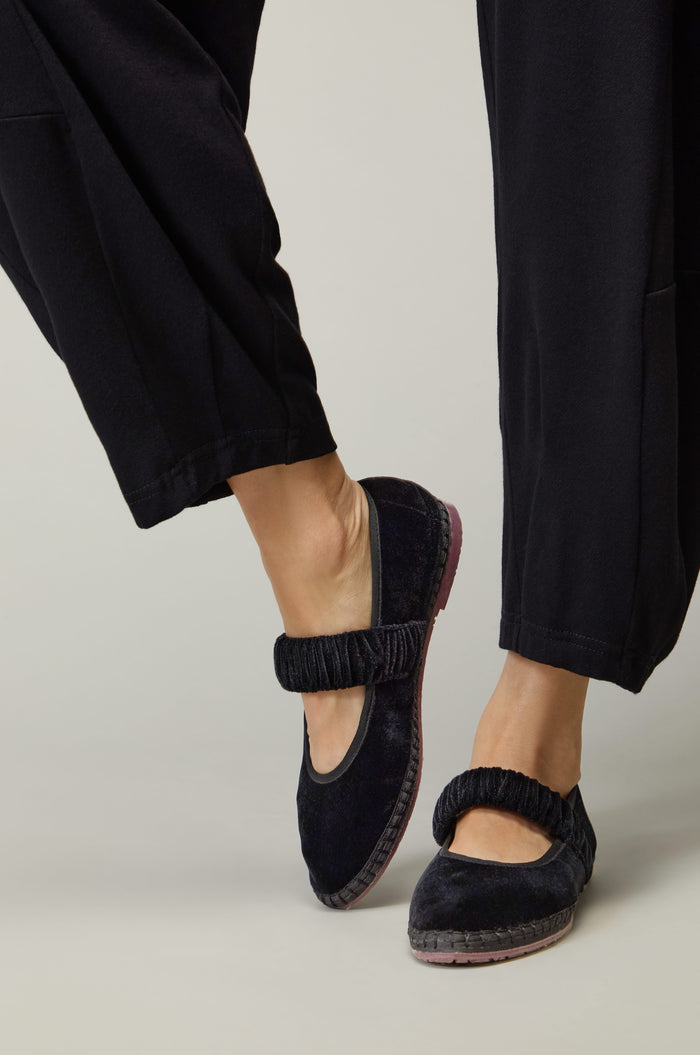 Wearing Mafalda Black Velvet Shoes—handmade in Spain with elastic straps and black velvet, embodying slow fashion—paired with black pants, one foot slightly raised.