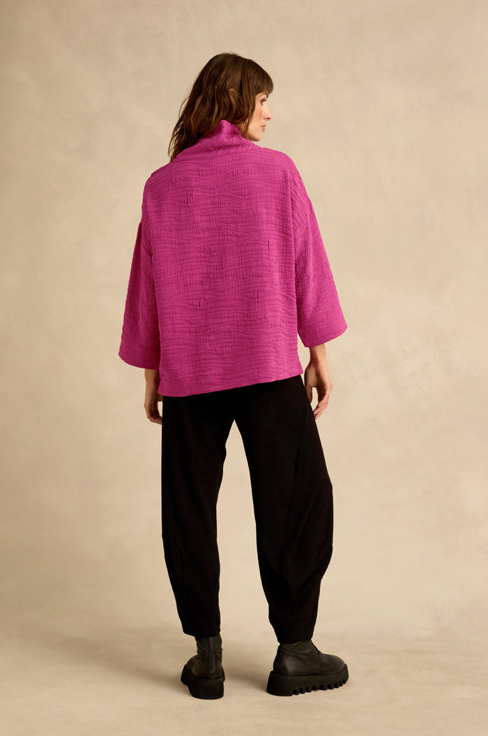 A person stands with their back turned, wearing the Magenta Wave Textured Crinkle Cowl Neck Top, along with loose black pants and black shoes, set against a neutral background.