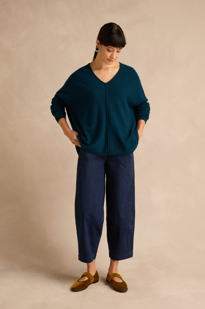 A woman stands against a beige background, wearing the Marine Merino Wool V Neck Jumper in teal, paired with dark blue wide-leg pants and brown loafers, her hands in her pockets as she looks down.