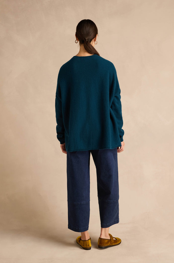 A person stands facing away against a neutral beige background, wearing the Marine Merino Wool V Neck Jumper in teal, paired with dark wide-leg pants and yellow loafers.