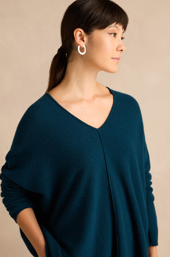 A woman wearing the Marine Merino Wool V Neck Jumper in teal, styled as a classic women's knit sweater with white hoop earrings, stands against a beige background and looks to the side.