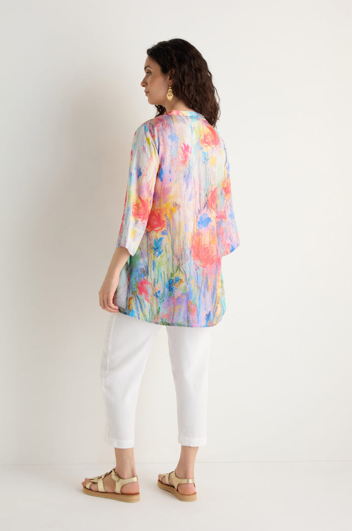 A woman faces away, modeling the Meadow Dreams Flared Shirt with a colorful floral print and Nehru collar, paired with white pants, beige sandals, and gold earrings against a plain white background.