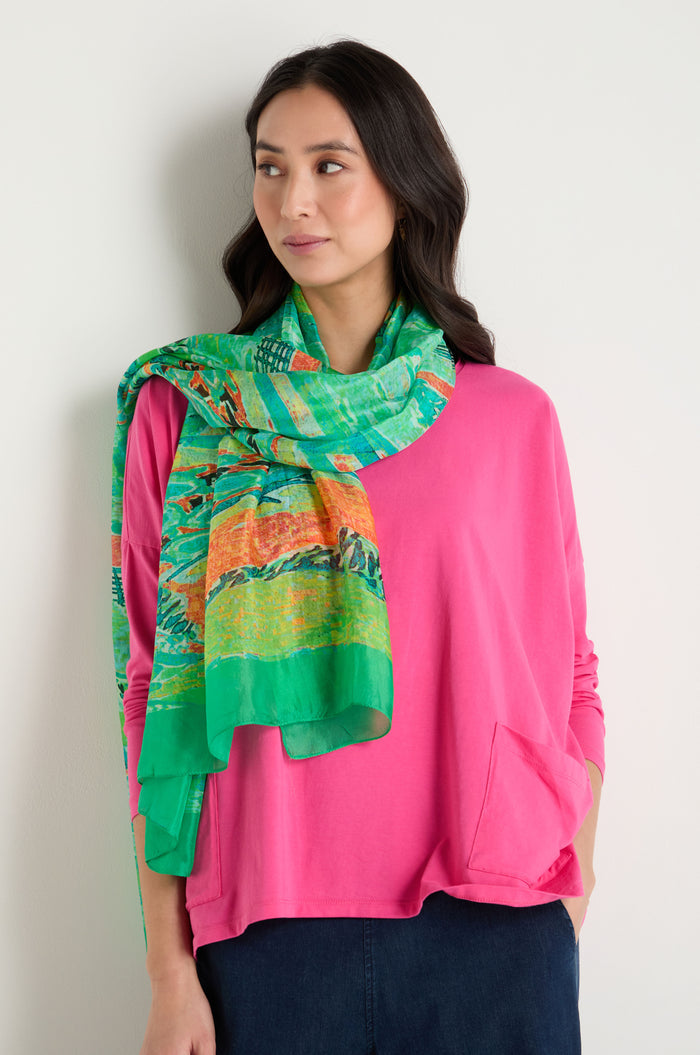 A woman stands against a plain light-colored wall, wearing a bright pink long-sleeve top and the Meadow Silk Scarf, a colorful pure silk accessory with an abstract print.