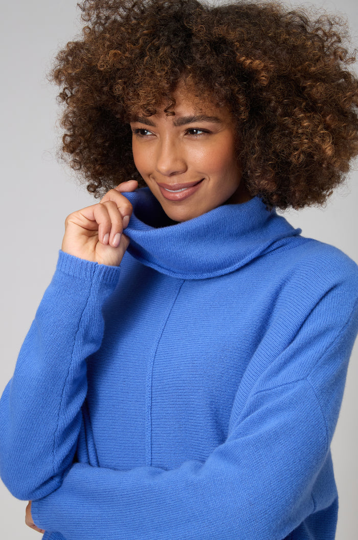 Person with curly hair wearing a blue Merino Cowl Neck Jumper, smiling slightly and touching the collar with one hand.
