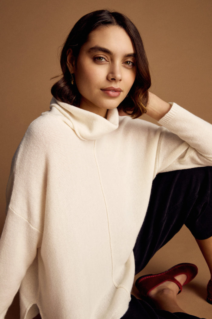 A woman with shoulder-length dark hair models the Merino Cowl Neck Jumper in cream and black pants with red shoes, seated against a brown background. She gazes confidently at the camera, showcasing luxurious women's knitwear.