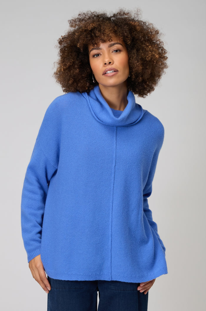 A woman with curly hair wears a loose-fitting, long-sleeve Merino Cowl Neck Jumper in blue and dark pants, standing against a plain light background.