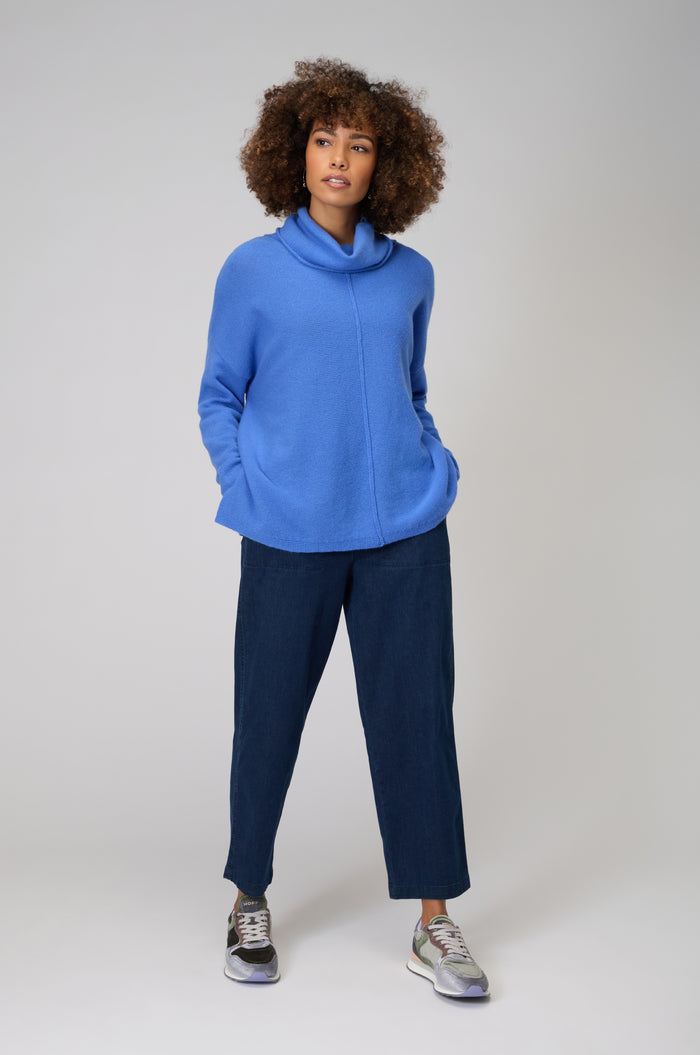 A woman stands facing forward with hands in her pockets, wearing a Merino Cowl Neck Jumper in blue, dark blue pants, and gray sneakers, against a plain light background.