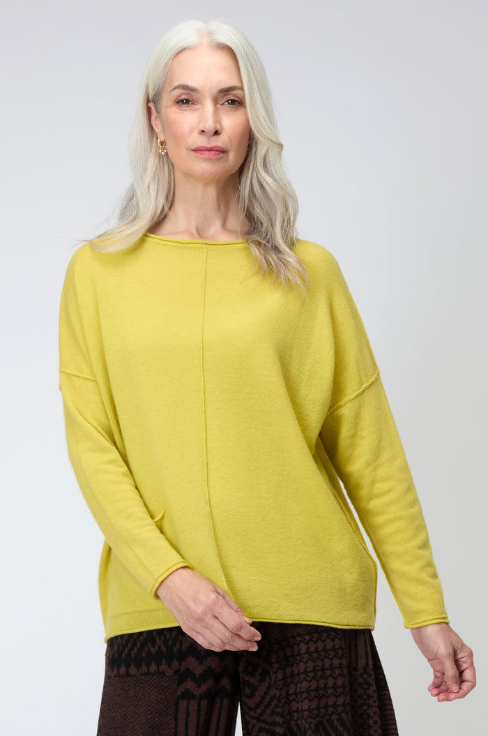 An older woman with long gray hair stands facing the camera against a plain background, wearing a yellow Merino Pocket Jumper and dark patterned pants.