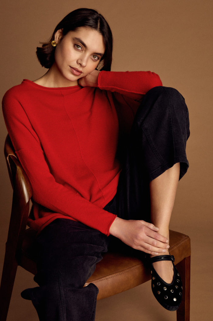 A woman with dark hair sits on a brown chair against a brown background, wearing the red Merino Pocket Jumper, dark pants, and black shoes, with one arm resting on her knee.