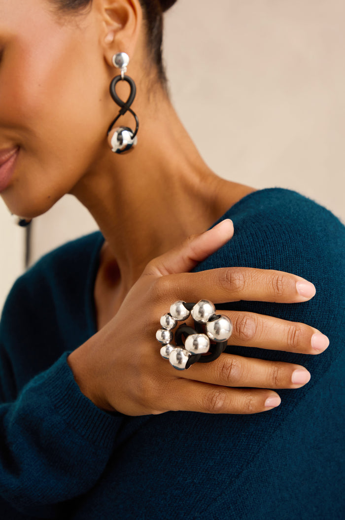 A woman wearing a teal sweater showcases the Metal Spheres Adjustable Ring—a large, sculptural silver statement piece adorned with spherical accents—along with matching handmade earrings, all crafted in France.