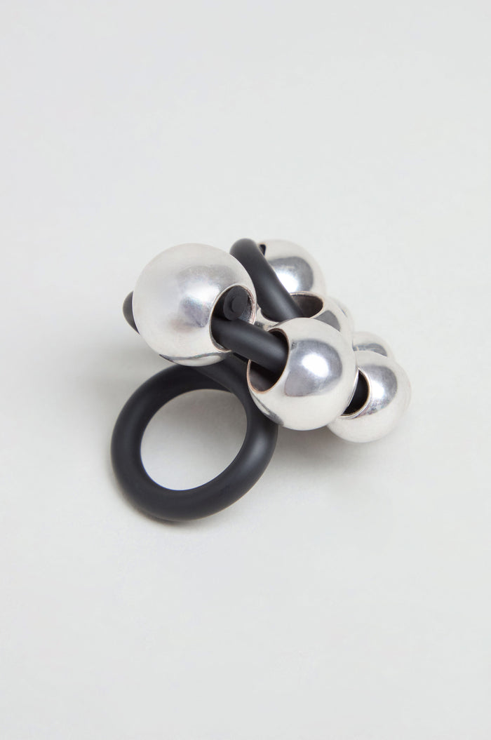 The Metal Spheres Adjustable Ring showcases a sculptural design with interconnected black and silver metallic spheres, elegantly displayed on a plain light background.