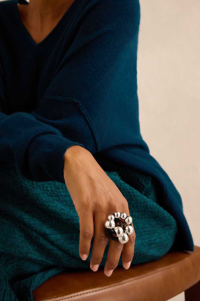 Wearing a blue sweater and teal skirt while seated on a brown chair, the person displays the Metal Spheres Adjustable Ring—a large, sculptural silver piece handmade in France—on their hand.