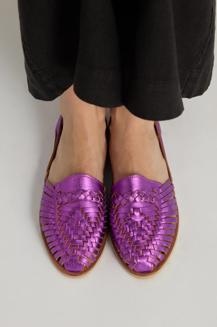 A person stands on a light surface wearing black pants and Metallic Purple Alegre Leather Huarache Sandals, handcrafted in Mexico.