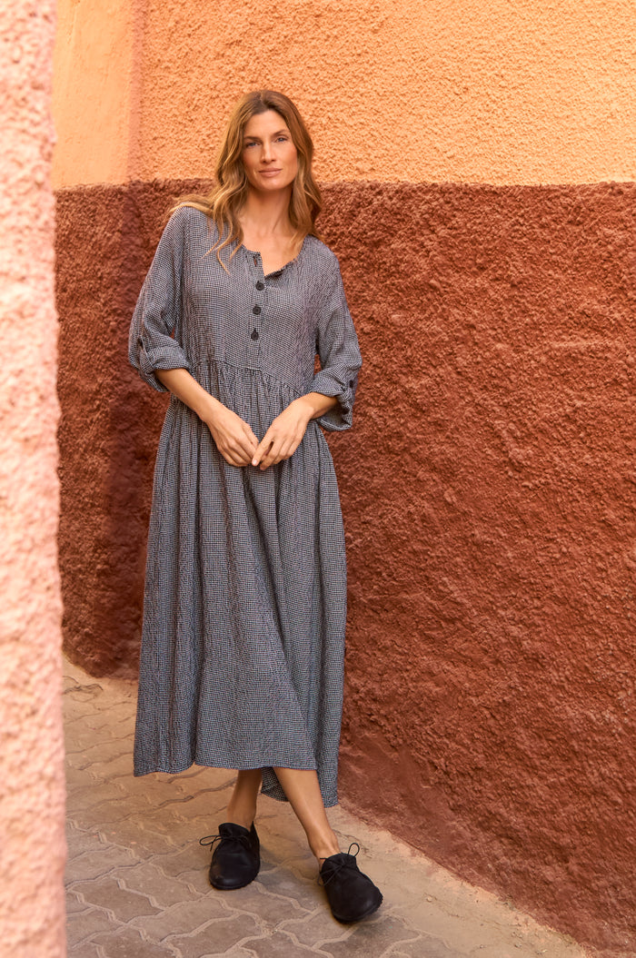 A woman stands against a textured red and peach wall, wearing the Micro Gingham Bubble Dress in gray with black buttons and black shoes.