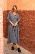 A woman stands against a textured red and peach wall, wearing the Micro Gingham Bubble Dress in gray with black buttons and black shoes.