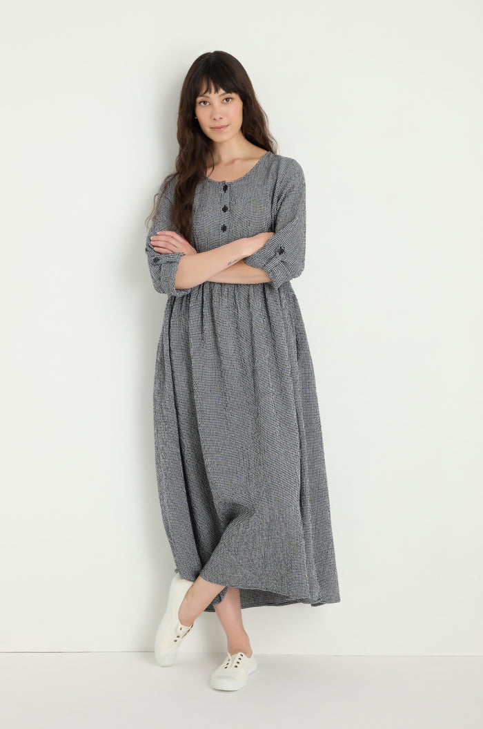 A woman with long dark hair stands against a white wall, arms crossed, wearing the Micro Gingham Bubble Dress in gray with buttons and white sneakers.