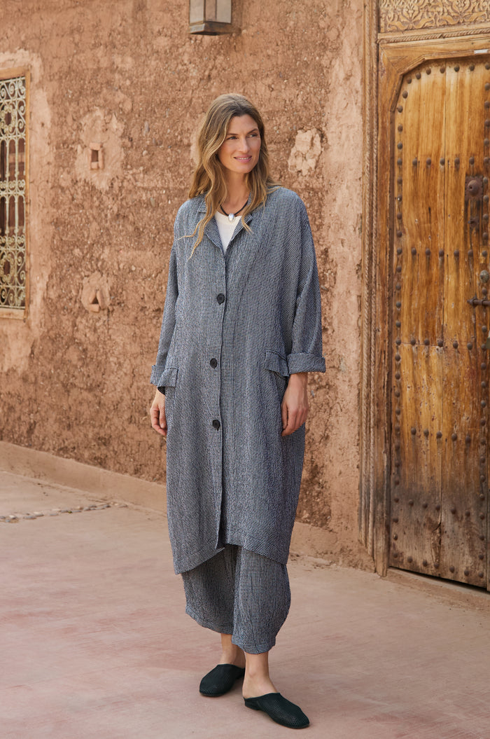 A woman stands outdoors by a textured wall and wooden door, wearing a long, loose-fitting gray Micro Gingham Boxy Coat with matching pants and black slip-on shoes.