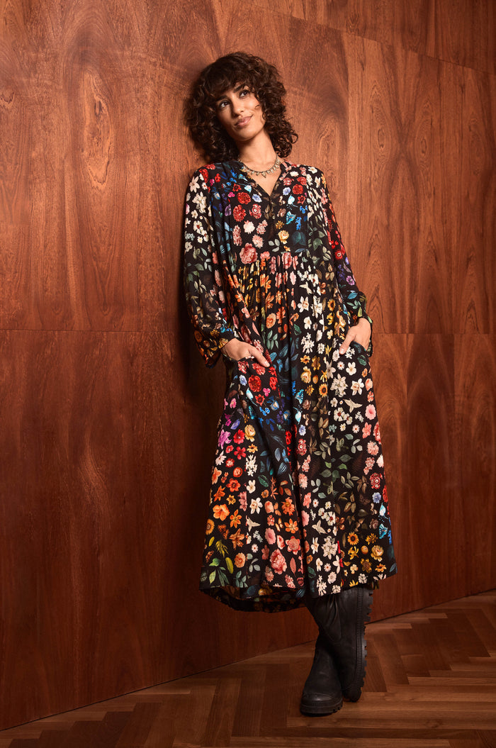 A person with curly hair stands against a wood-paneled wall, wearing the Midnight Garden Viscose Dress—a long black floral print dress with colorful patterns—paired with black boots and hands in pockets, on a wooden floor.