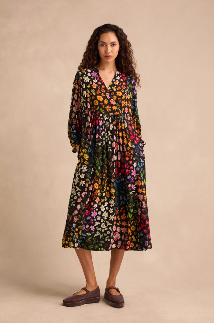 A person stands against a beige background wearing the Midnight Garden Viscose Dress, a long-sleeve, ankle-length black floral crepe dress with a colorful print, paired with brown shoes.