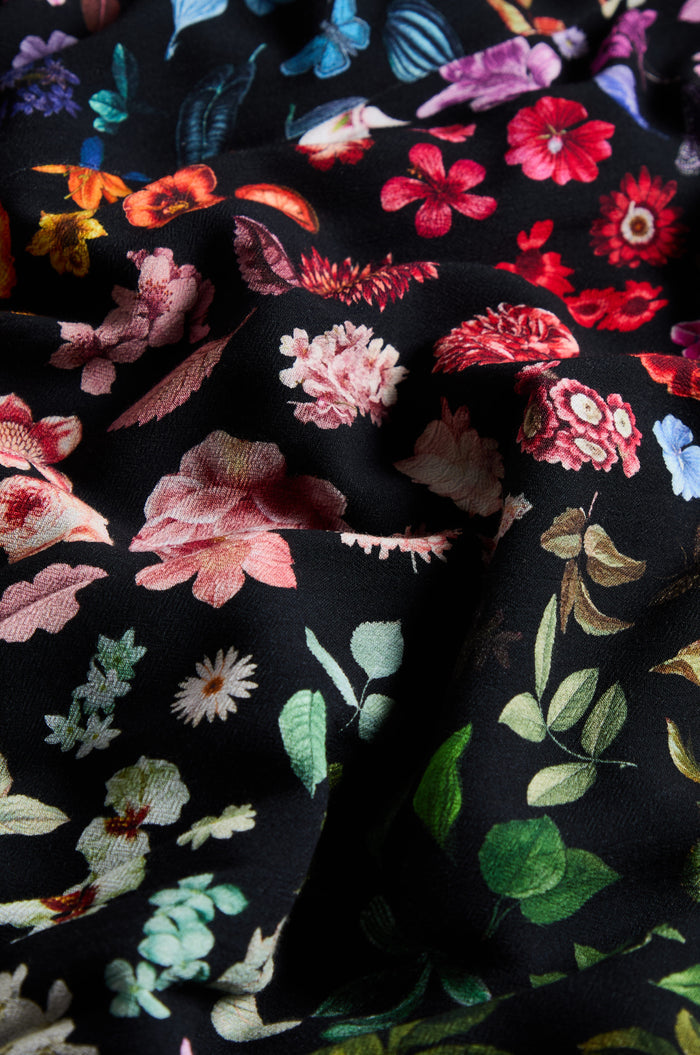 Close-up of the Midnight Garden Viscose Dress showcases black fabric adorned with colorful printed flowers and leaves, blending vibrant reds, pinks, greens, and blues on a dark background—ideal for crafting an elegant floral crepe dress or a stylish wrap front dress.