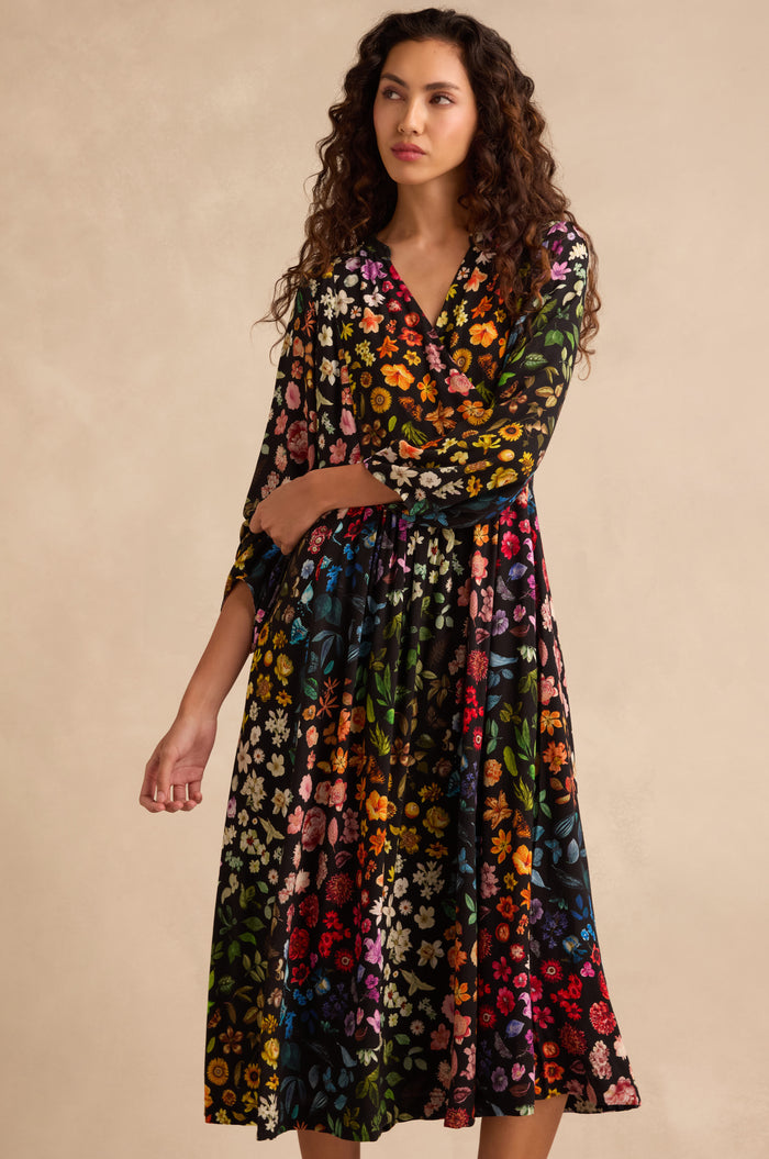 A woman wears the Midnight Garden Viscose Dress, featuring long sleeves, a vibrant floral print on black crepe fabric, and an elegant wrap-front design, as she stands against a beige background.