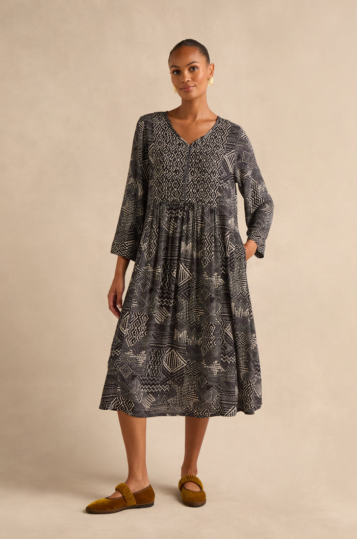 A woman stands against a beige background wearing the Mono Patchwork Ikat Dress, an ankle-length, long-sleeved A-line dress featuring white geometric patterns, paired with brown flat shoes.