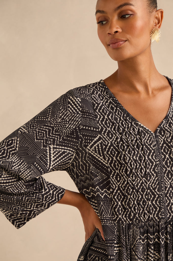 A woman wearing the Mono Patchwork Ikat Dress—a long-sleeve, black and white geometric A-line dress—paired with gold sunburst earrings and her hair pulled back, stands against a neutral background.