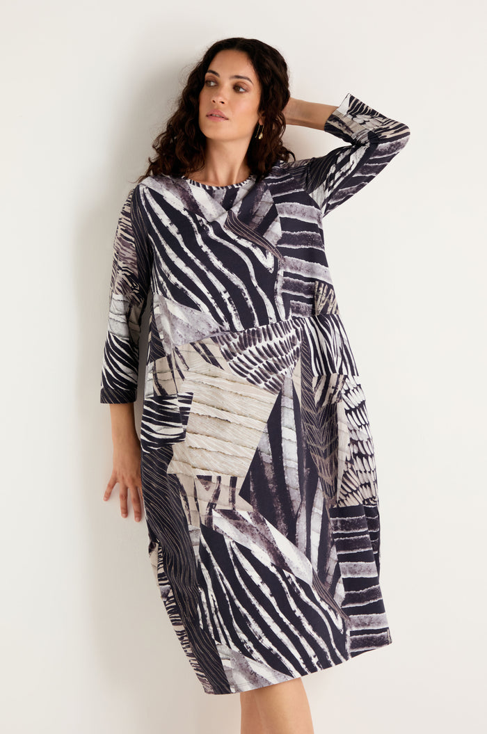 A woman wearing the Monochrome Patchwork Jersey Dress—a loose-fitting, long-sleeve dress featuring an abstract geometric and striped pattern in black, white, and beige—stands against a plain light-colored wall.