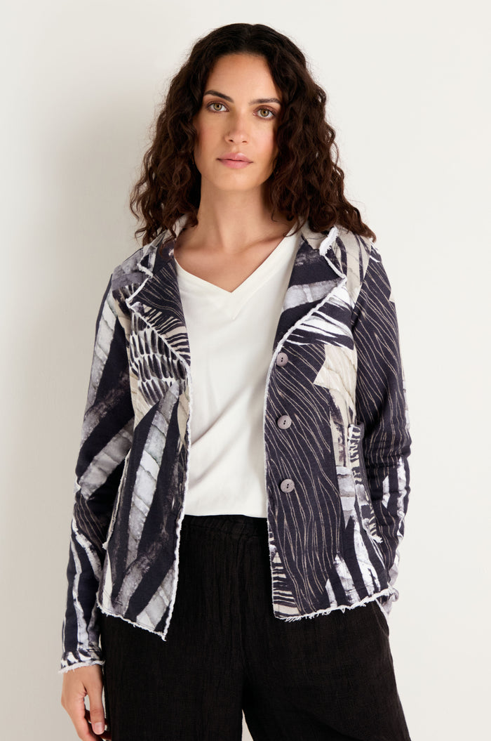A woman with long curly hair stands against a plain light background wearing the Monochrome Patchwork Jersey Jacket over a white cotton top and black pants.