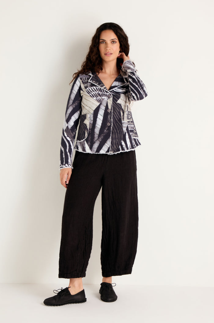 A woman stands against a plain wall, wearing the Monochrome Patchwork Jersey Jacket, paired with black wide-leg pants and black shoes, with one hand touching her hair.