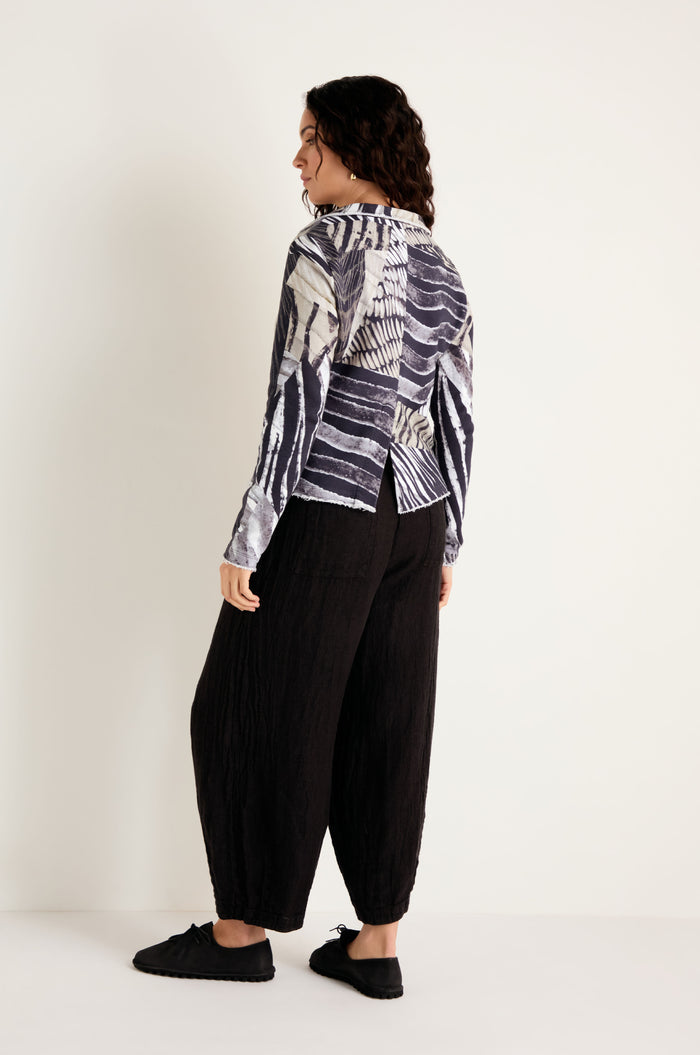 A person stands against a plain background, facing away from the camera, wearing a patterned long-sleeve shirt and loose black pants paired with black shoes; their outfit is enhanced by the Monochrome Patchwork Jersey Jacket, which adds an extra layer of texture.
