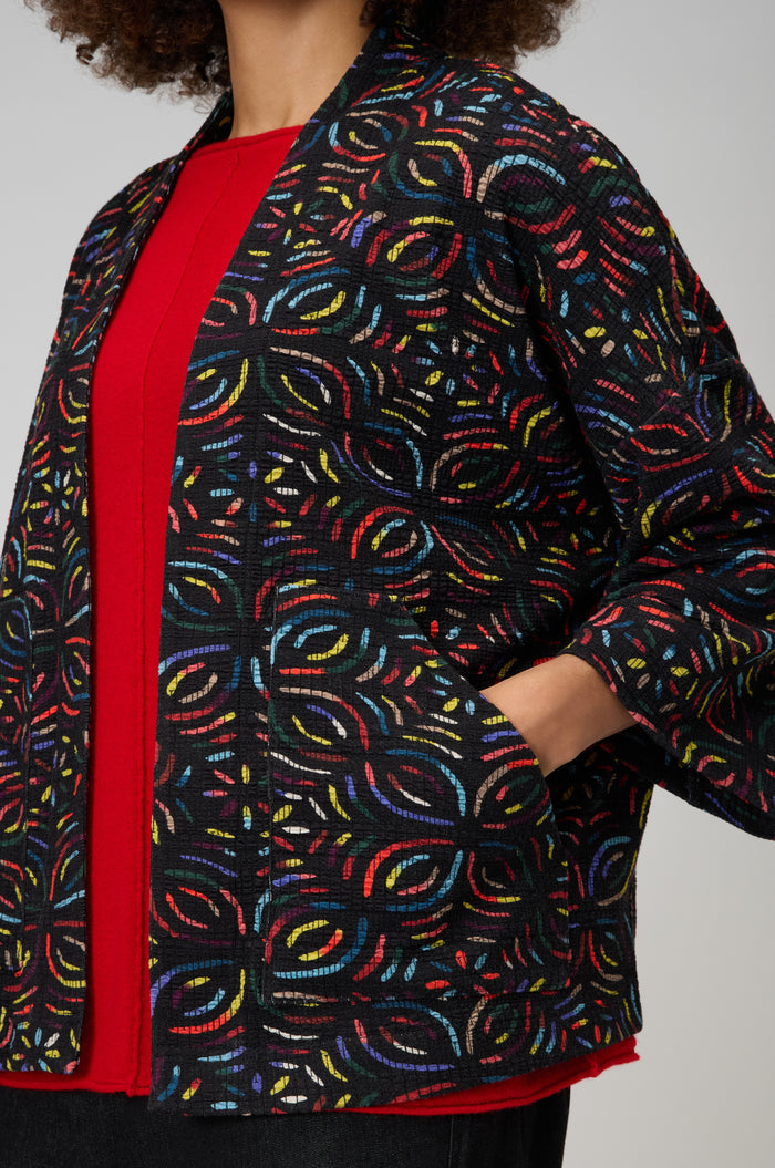 A person wearing a red top and the Mosaic Jacquard Jersey Jacket with colorful embroidered patterns stands with one hand in their pocket.
