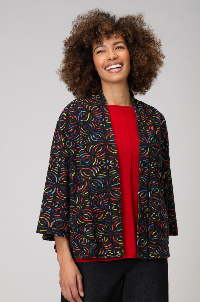 A woman smiles, wearing the Mosaic Jacquard Jersey Jacket with colorful circular patterns over a red top and dark pants, standing against a plain light background.