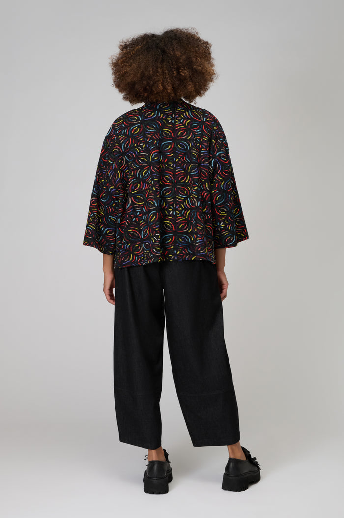 Seen from behind, the person wears the Mosaic Jacquard Jersey Jacket with a colorful swirl pattern, paired with wide-leg black pants and black platform shoes. The look is modern and effortlessly stylish against a plain background.