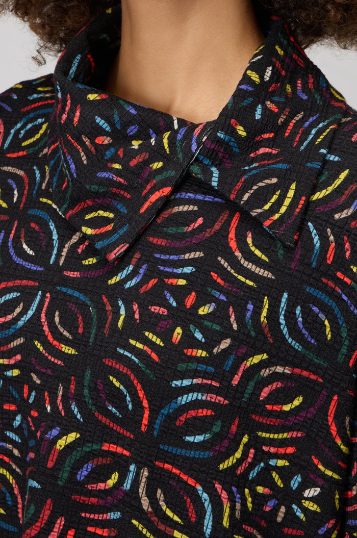 Close-up of a person's upper chest and neck wearing the Mosaic Jacquard Jersey Top, a black collared jersey with a colorful abstract circular pattern in a relaxed fit.