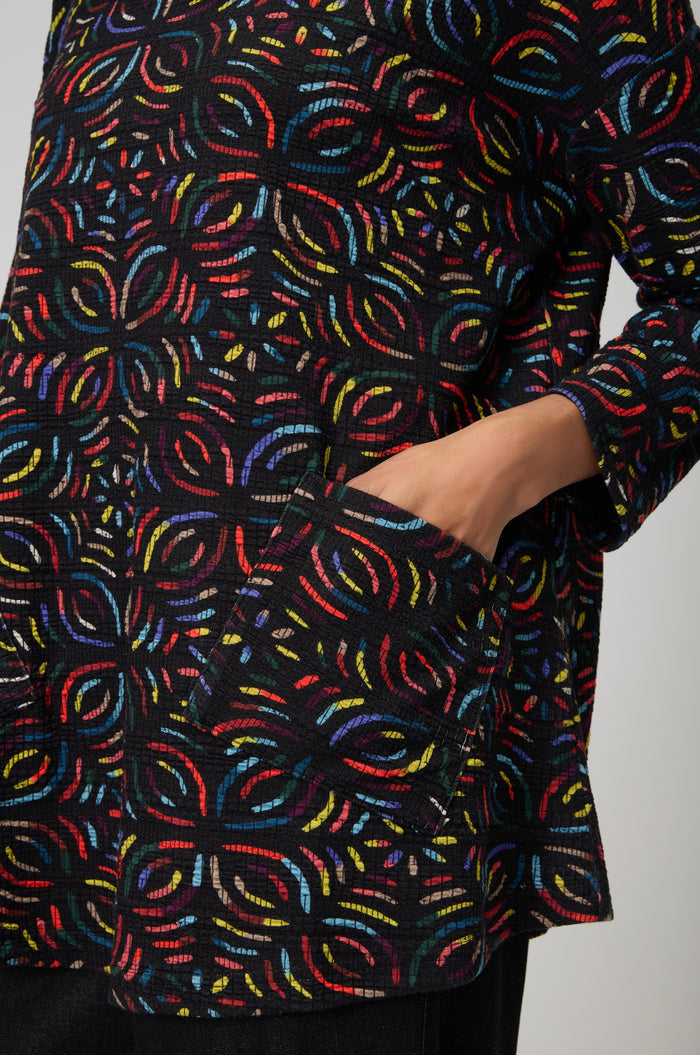 A person wears the Mosaic Jacquard Jersey Top—a relaxed fit black jersey with a colorful geometric pattern—and places their right hand in a side pocket.