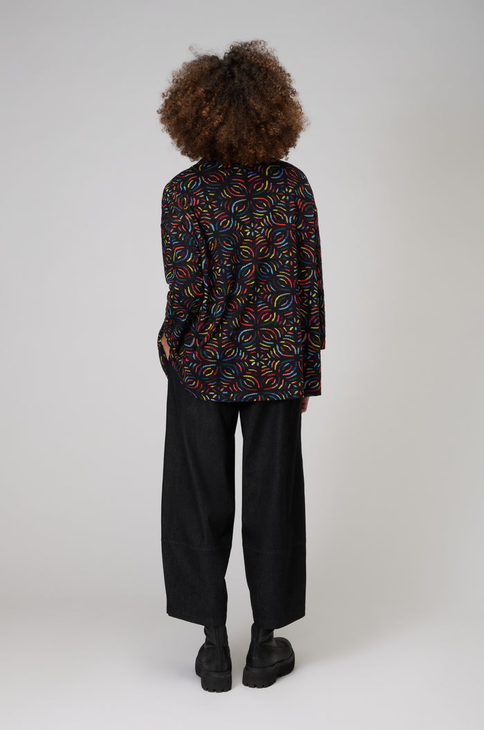 A person with curly hair faces away, wearing the Mosaic Jacquard Jersey Top with a colorful mosaic pattern, paired with loose black pants and black shoes, set against a plain background.