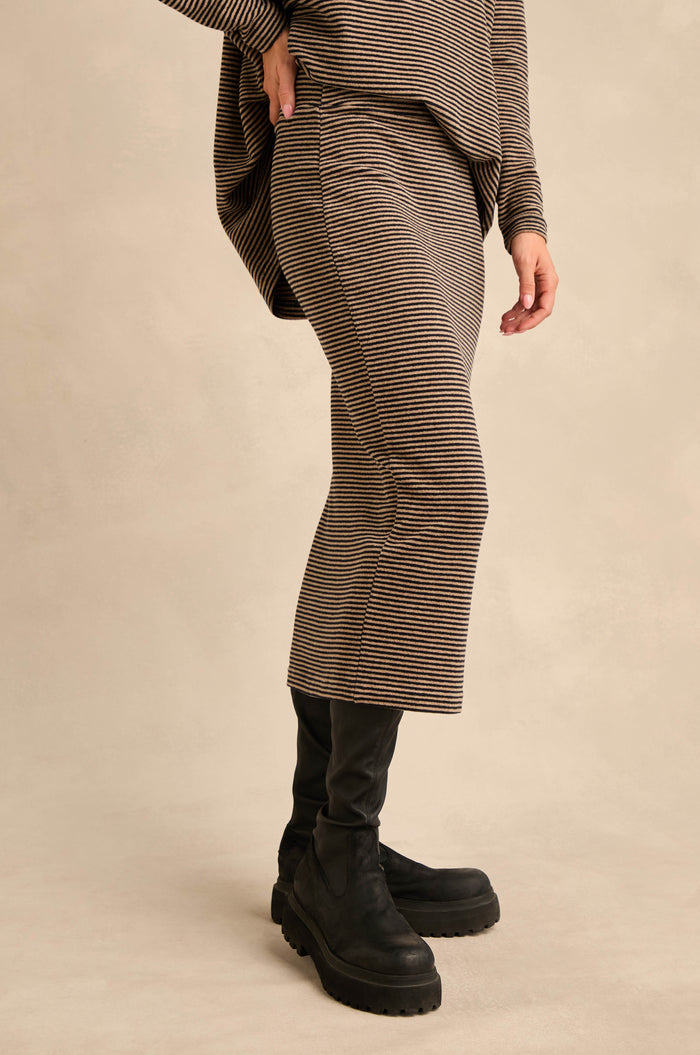 A person wearing the Narrow Stripe Jersey Tube Skirt in brown and black narrow stripes, paired with a long matching top and black boots, stands against a plain beige background.
