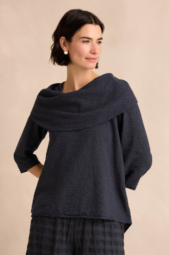 A woman models the Navy Draped Neck Top, featuring a wide cowl neck, three-quarter sleeves, and a textured design. Crafted from soft cotton-viscose jersey for a relaxed fit, she stands against a neutral background.