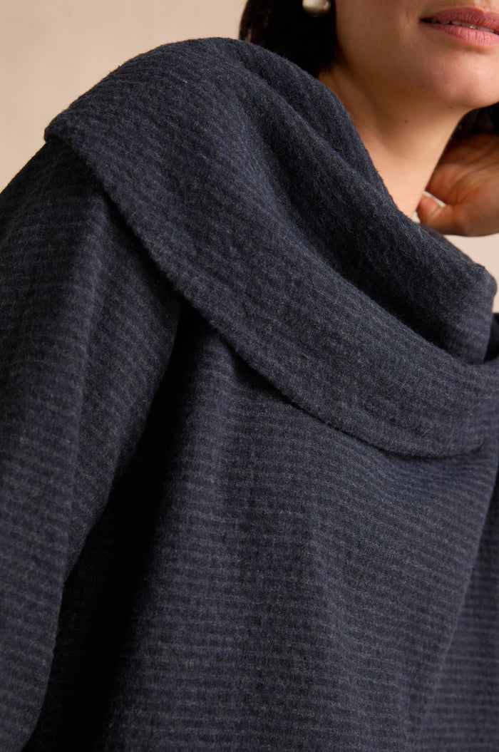 Close-up of a person wearing the Navy Draped Neck Top, featuring a textured dark navy hue and a wide, folded draped collar. The relaxed fit accentuates the soft structure of its cotton-viscose jersey fabric. Part of the face and hand are visible.