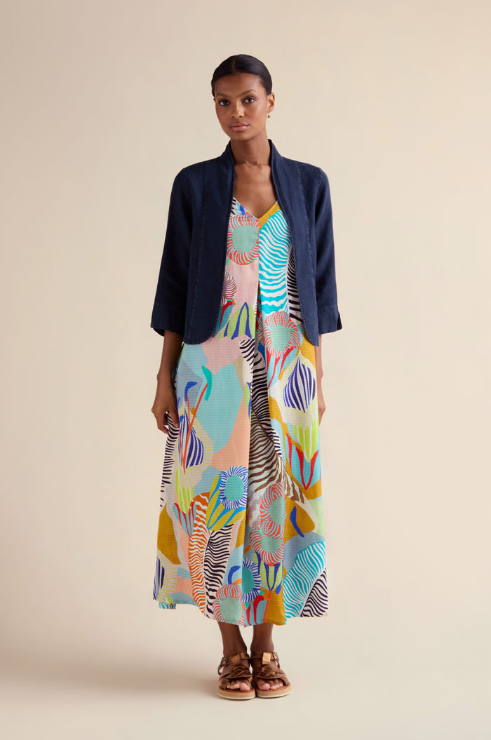 A woman styles the Navy Linen Cropped Jacket over a vibrant, abstract-print maxi dress and tan sandals against a neutral background.