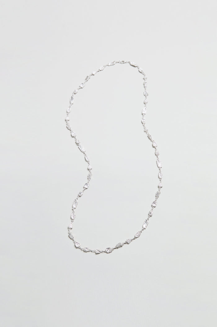 The Nugget Chain Necklace, featuring small, twisted silver links, is elegantly arranged in an S-shape on a plain, textured surface.