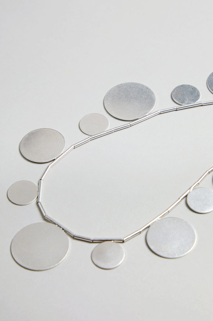 The Orbit Disc Necklace, featuring flat, round silver discs in assorted sizes, is displayed on a light grey background.