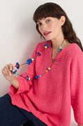 A woman in a textured pink top and dark pants holds the Orbit Necklace—an abstract, colorful, geometric accessory—while sitting against a plain background.