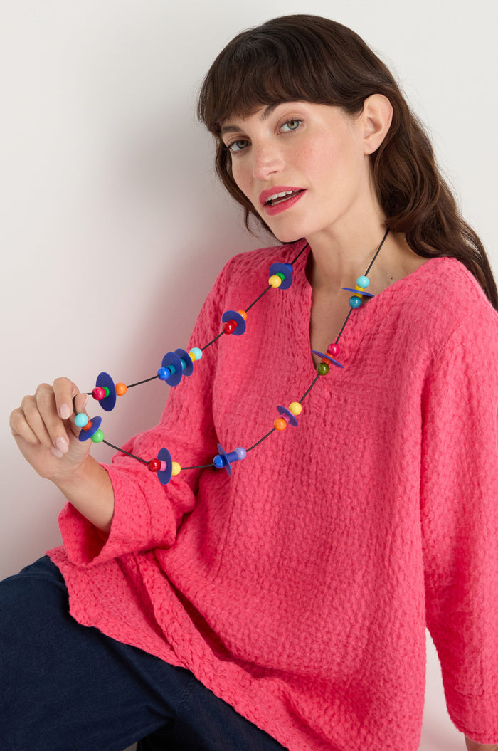 A woman in a textured pink top and dark pants holds the Orbit Necklace—an abstract, colorful, geometric accessory—while sitting against a plain background.