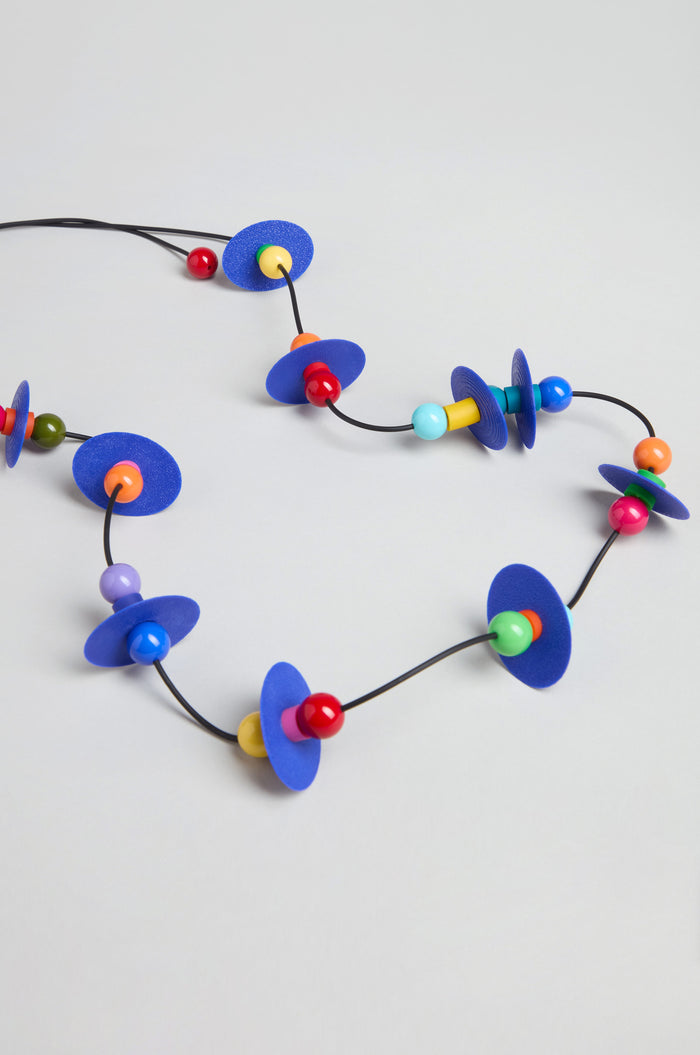 The Orbit Necklace showcases abstract blue flat discs and multicolored round beads on a black cord, arranged against a light gray background.