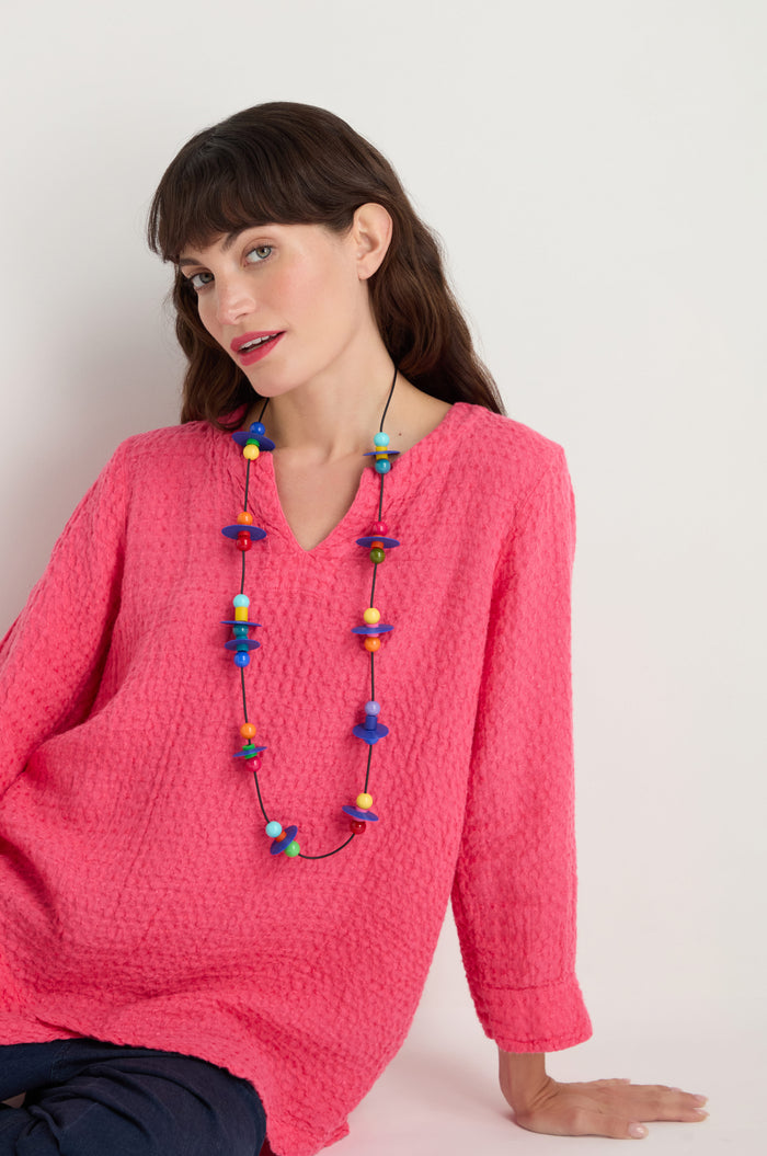 A woman in a textured pink top, dark pants, and the Orbit Necklace with an abstract geometric design sits against a white background.