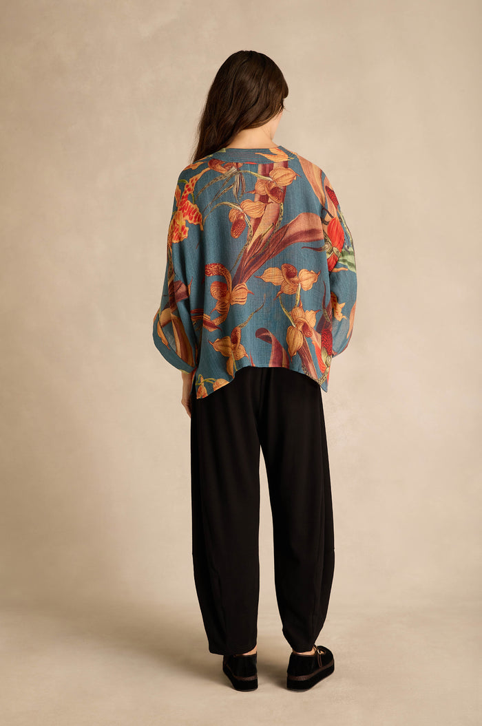 A person stands with their back to the camera, wearing the Orchid Wool Kimono—a loose, long-sleeved top adorned with an oversized slipper orchid print—paired with black pants and black shoes, set against a neutral background.