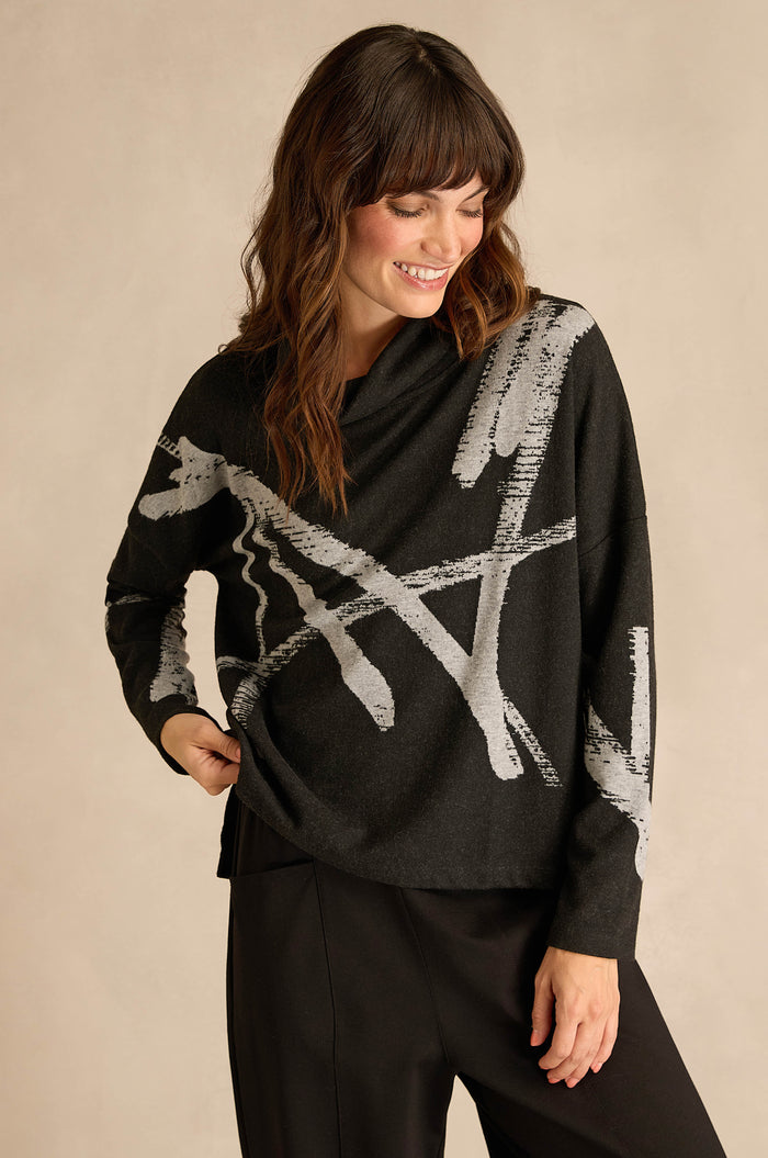 A woman wearing the Paint Stripes Long Sleeve Top—a relaxed fit, black jersey adorned with gray abstract brushstroke patterns—paired with black pants, stands against a beige background.